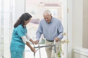 A home healthcare worker helping a senior man with a walker walking through the front door of his house.